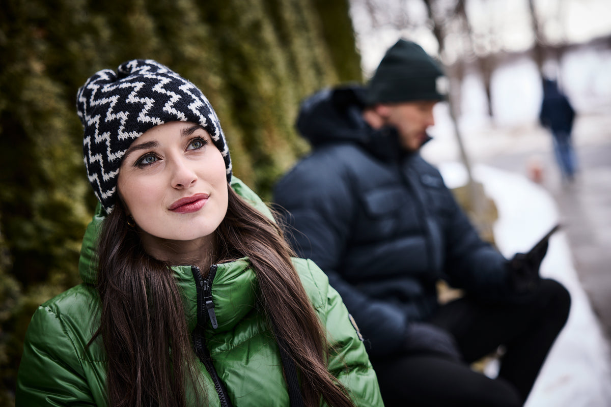 Woman wearing a green jacket and patterned beanie sitting outdoors with a blurred person in the background.